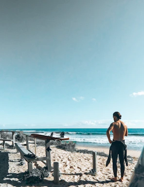 A person with a wetsuit tied around their waist stands on a sandy beach, looking towards the ocean. A surfboard is placed on a wooden bench next to them. The sky is clear and blue, creating a peaceful coastal scene.