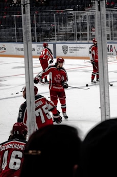Players dressed in red hockey uniforms are on an ice rink, actively engaging in what appears to be a practice or game. The background shows empty rows of seats and glass panels surrounding the rink.