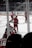 Players dressed in red hockey uniforms are on an ice rink, actively engaging in what appears to be a practice or game. The background shows empty rows of seats and glass panels surrounding the rink.