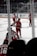 Players dressed in red hockey uniforms are on an ice rink, actively engaging in what appears to be a practice or game. The background shows empty rows of seats and glass panels surrounding the rink.