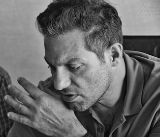 A black and white close-up portrait of a man lost in thought, his gaze directed downward. His right hand rests lightly on his left shoulder, capturing the texture of his stubble and the subtle expression of contemplation.