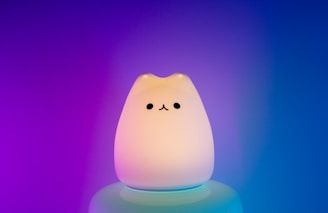 A cute, cat-shaped night light emits a soft glow with a gradient of purple and blue in the background. The lamp has simple black eyes and a tiny nose, creating an adorable and minimalist face.