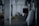 A white dog stands in the middle of a dimly lit barn aisle. The barn features concrete walls and pillars that stretch down the corridor, with hay scattered on the floor. Large sacks and a dark red horse blanket are visible to the side, indicating an equestrian setting.