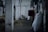 A white dog stands in the middle of a dimly lit barn aisle. The barn features concrete walls and pillars that stretch down the corridor, with hay scattered on the floor. Large sacks and a dark red horse blanket are visible to the side, indicating an equestrian setting.