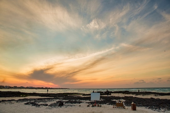 A serene beach scene at sunset with a dramatic sky painted in hues of orange, pink, and blue. The foreground features a sandy beach with scattered rocks and a few people walking. There is a small setup with wooden boxes and a sign that reads 'LOVE' nearby.