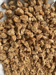 A collection of irregularly shaped brown solid chunks, likely some form of sweet or food item, with a grainy texture. The chunks are piled on a light-colored surface.