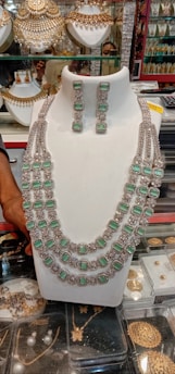 A display of ornate jewelry featuring a large necklace adorned with multiple strands and green gemstones, paired with matching earrings. The background shows various other jewelry items such as gold embellishments and decorative arrangements. A hand can be seen adjusting or holding the display piece.
