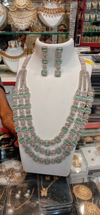 A display of ornate jewelry featuring a large necklace adorned with multiple strands and green gemstones, paired with matching earrings. The background shows various other jewelry items such as gold embellishments and decorative arrangements. A hand can be seen adjusting or holding the display piece.