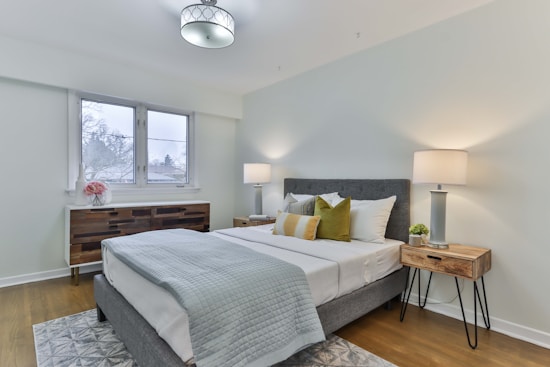 A modern bedroom with a neatly made bed featuring gray and white bedding accented with yellow and green pillows. Two wooden nightstands with lamps and a plant as decor sit on either side of the bed. A wooden dresser with decorative vases and a flower bouquet is positioned against the wall under a window. The room has soft lighting, wooden flooring, and a subtle rug under the bed.