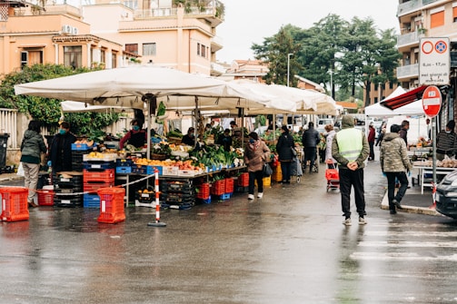 A bustling outdoor market scene with multiple stalls selling fresh fruits and vegetables under large white umbrellas. People are browsing and purchasing goods, and the area is lined with red and blue crates. Buildings and greenery are visible in the background, and the scene appears to have a damp ground indicating recent rain.