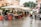 A bustling outdoor market scene with multiple stalls selling fresh fruits and vegetables under large white umbrellas. People are browsing and purchasing goods, and the area is lined with red and blue crates. Buildings and greenery are visible in the background, and the scene appears to have a damp ground indicating recent rain.