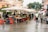 A bustling outdoor market scene with multiple stalls selling fresh fruits and vegetables under large white umbrellas. People are browsing and purchasing goods, and the area is lined with red and blue crates. Buildings and greenery are visible in the background, and the scene appears to have a damp ground indicating recent rain.