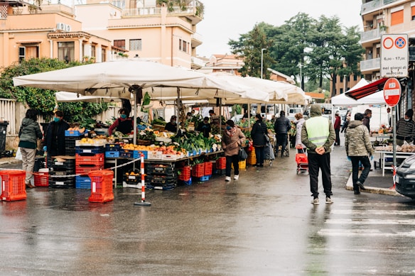 A bustling outdoor market scene with multiple stalls selling fresh fruits and vegetables under large white umbrellas. People are browsing and purchasing goods, and the area is lined with red and blue crates. Buildings and greenery are visible in the background, and the scene appears to have a damp ground indicating recent rain.