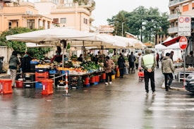 A bustling outdoor market scene with multiple stalls selling fresh fruits and vegetables under large white umbrellas. People are browsing and purchasing goods, and the area is lined with red and blue crates. Buildings and greenery are visible in the background, and the scene appears to have a damp ground indicating recent rain.