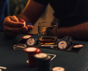A hand holding several poker chips, with a glass of whiskey and a stack of poker chips on a table. The setting appears to be a dimly lit environment, suggesting an intimate or focused atmosphere.