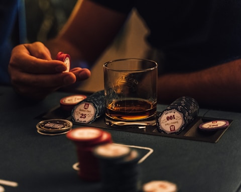 A hand holding several poker chips, with a glass of whiskey and a stack of poker chips on a table. The setting appears to be a dimly lit environment, suggesting an intimate or focused atmosphere.