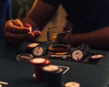 A hand holding several poker chips, with a glass of whiskey and a stack of poker chips on a table. The setting appears to be a dimly lit environment, suggesting an intimate or focused atmosphere.