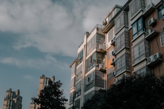 A residential building with multiple floors and a modern architectural design. The structure is characterized by large windows covered with metal bars, air conditioning units attached to several windows, and balconies. Sunlight casts a warm glow on one side of the building, creating a contrast between light and shadow. Trees partially obscure the lower part of the view, with additional buildings visible in the background under a cloudy sky.
