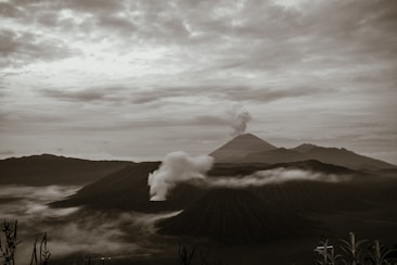 A dramatic landscape featuring a series of mist-covered volcanoes, with one actively emitting smoke. The sky is overcast with thick clouds, adding a moody atmosphere. Sparse vegetation frames the foreground, enhancing the natural setting.