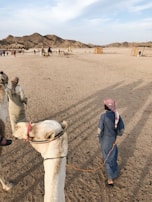 A sandy desert landscape with a few people and camels visible. A person wearing traditional attire leads a camel, while other people and camels can be seen in the distance near some simple structures. The background features rugged hills under a partly cloudy sky.