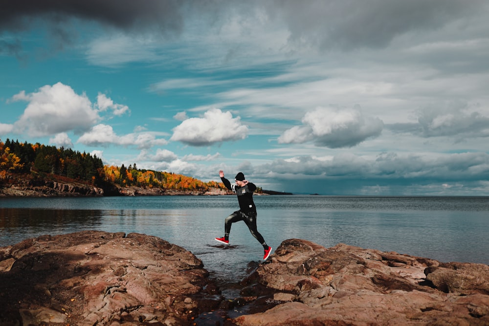 man in black jacket and black pants sitting on brown rock near body of water during