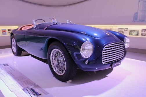 A vintage, elegant blue sports car with a classic design is displayed in a well-lit showroom. It features a sleek body, large round headlights, and a prominent front grille. The car is positioned on a glossy white platform with a few display plaques nearby. The background includes softly lit walls showcasing framed photographs or posters.