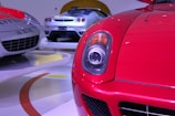 A close-up view of a bright red sports car headlight with a sleek aerodynamic design. In the background, two other high-end sports cars with silver and white bodies are displayed on a polished showroom floor.