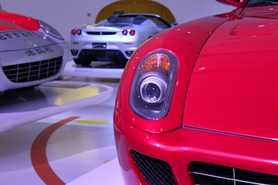 A close-up view of a bright red sports car headlight with a sleek aerodynamic design. In the background, two other high-end sports cars with silver and white bodies are displayed on a polished showroom floor.