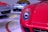 A close-up view of a bright red sports car headlight with a sleek aerodynamic design. In the background, two other high-end sports cars with silver and white bodies are displayed on a polished showroom floor.