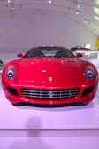 A bright red sports car is displayed indoors, positioned on a white platform with a modern, curved ceiling. The car features a sleek, aerodynamic design with prominent headlights, a polished front grille, and a distinct logo on the hood.