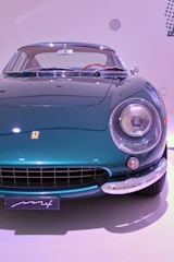 A vintage sports car with a sleek chrome bumper and a shiny teal exterior. The front headlights are prominent, and the grille has vertical slats. The car’s body reflects light, indicating a well-polished surface, and it is positioned against a simple white backdrop.