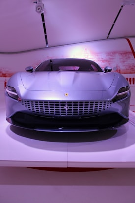 A sleek and modern silver sports car with a distinctive front grille and aerodynamic design is positioned in an indoor setting with white and red background elements. The headlights are narrow, and the car bears a logo in the center of the grille. Overhead lighting highlights the car's contours.