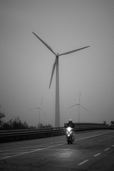 A black and white image with a person riding a motorcycle on a road. In the background, three large wind turbines tower over the scene. The setting appears to be overcast, contributing to a moody and serene atmosphere.