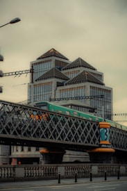 An urban scene featuring a train traveling on a lattice-style bridge over a city street, with modern buildings in the background. The structures have triangular roof designs, and the bridge shows vibrant artwork on the supporting pillars.