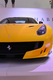 A vibrant yellow sports car is displayed prominently, featuring sleek design elements like a low front profile, aerodynamic curves, and a prominent logo on the grille. The background is minimalistic with parts of letters visible, hinting at a possible exhibition or event space.