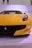 A vibrant yellow sports car is displayed prominently, featuring sleek design elements like a low front profile, aerodynamic curves, and a prominent logo on the grille. The background is minimalistic with parts of letters visible, hinting at a possible exhibition or event space.