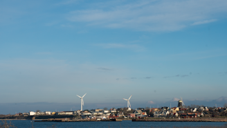 A coastal town with several wind turbines in the background. The area includes a mix of residential buildings that line the shore, with a prominent windmill visible to the right. The sky is clear with a few clouds, and the sea in the foreground is calm.