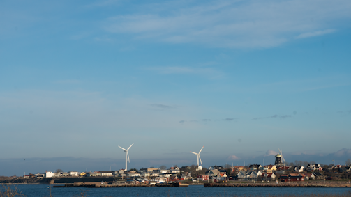 A coastal town with several wind turbines in the background. The area includes a mix of residential buildings that line the shore, with a prominent windmill visible to the right. The sky is clear with a few clouds, and the sea in the foreground is calm.