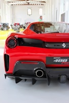 A red sports car is displayed in a bright showroom, featuring a sleek rear design with a dual exhaust system. The room has an array of automotive parts lined up in the background with large windows providing natural lighting. A person is visible standing in the background, wearing a green shirt.
