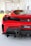A red sports car is displayed in a bright showroom, featuring a sleek rear design with a dual exhaust system. The room has an array of automotive parts lined up in the background with large windows providing natural lighting. A person is visible standing in the background, wearing a green shirt.