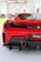 A red sports car is displayed in a bright showroom, featuring a sleek rear design with a dual exhaust system. The room has an array of automotive parts lined up in the background with large windows providing natural lighting. A person is visible standing in the background, wearing a green shirt.