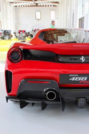 A red sports car is displayed in a bright showroom, featuring a sleek rear design with a dual exhaust system. The room has an array of automotive parts lined up in the background with large windows providing natural lighting. A person is visible standing in the background, wearing a green shirt.