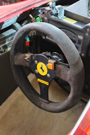 A close-up view of a racing car's steering wheel. The steering wheel is by Momo and features a prominent emblem with a black prancing horse on a yellow background. There are various buttons and switches around the steering wheel, including indications for neutral and off positions. The dashboard includes gauges and a branding sticker from Magneti Marelli.