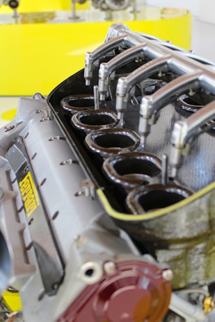 A close-up view of a high-performance engine, featuring multiple intake manifolds made of polished metal, partially unscrewed, revealing inner components. The engine has a sturdy, industrial design with a visible logo, set against a bright yellow background that highlights its mechanical complexity.