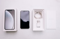 A smartphone package lies open, revealing a black smartphone, a white charging cable, a wall adapter, and the packaging box with the phone's image on the cover. The background is a plain, light-colored surface.
