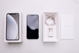 A smartphone package lies open, revealing a black smartphone, a white charging cable, a wall adapter, and the packaging box with the phone's image on the cover. The background is a plain, light-colored surface.