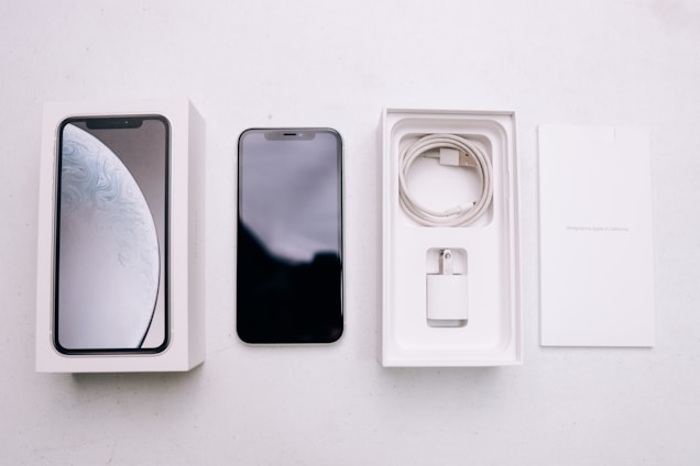 A smartphone package lies open, revealing a black smartphone, a white charging cable, a wall adapter, and the packaging box with the phone's image on the cover. The background is a plain, light-colored surface.