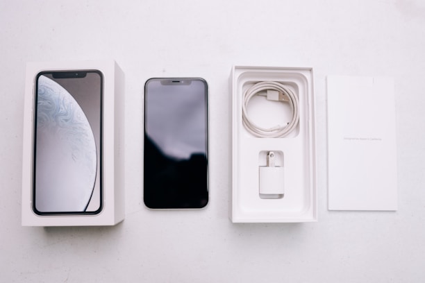 A smartphone package lies open, revealing a black smartphone, a white charging cable, a wall adapter, and the packaging box with the phone's image on the cover. The background is a plain, light-colored surface.