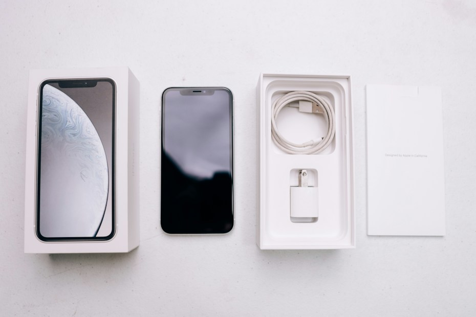 A smartphone package lies open, revealing a black smartphone, a white charging cable, a wall adapter, and the packaging box with the phone's image on the cover. The background is a plain, light-colored surface.