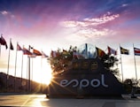 A sculpture with the logo 'ESPOL' is surrounded by numerous international flags. The sun is setting behind the sculpture, casting warm light and shadows across the scene. A clear sky with some clouds is visible in the background.